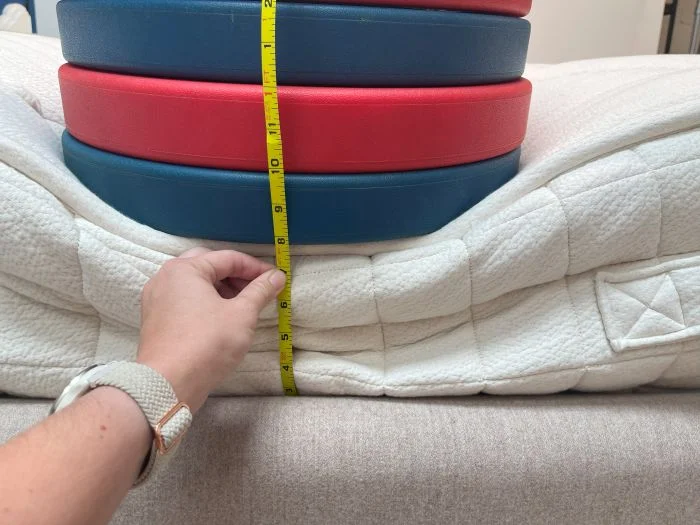 A stack of weight plates sit on top of the Avocado Luxury Organic mattress. A hand is measuring the sinkage with a tape measure.