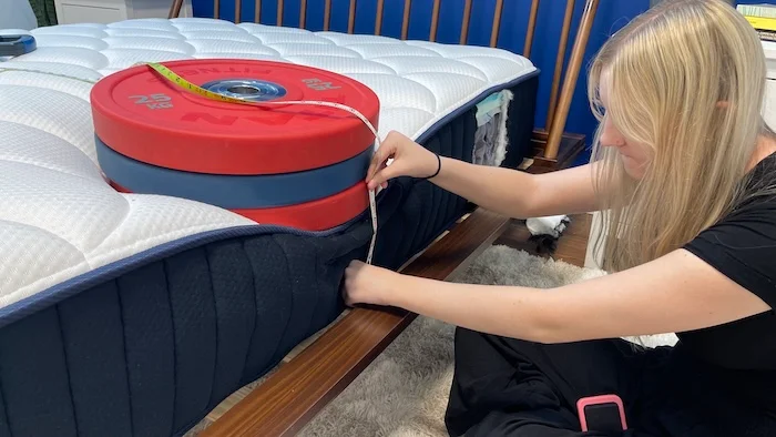 A woman kneeling next to the DreamCloud mattress. A stack of weights is resting on the bed's surface and the woman is measuring the sinkage with a tape measure.