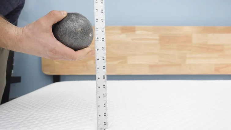 A man holding a steel ball and yardstick above a mattress, about to measure its sinkage.