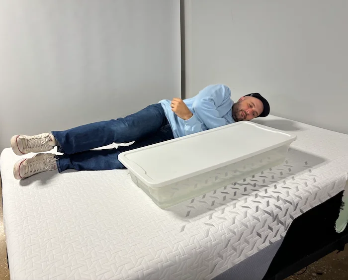 A man lying on the Nectar Premier Memory Foam mattress next to a plastic bin of water. He's rolling back and forth to see how much the water moves.