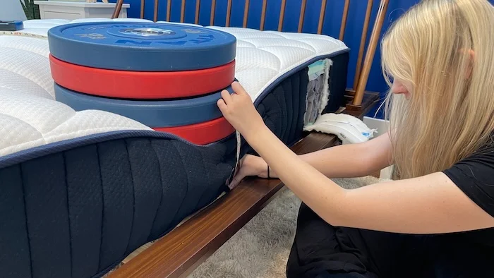 A woman kneeling next to the DreamCloud mattress. A stack of weights is resting on the bed's surface and the woman is measuring the sinkage with a tape measure.