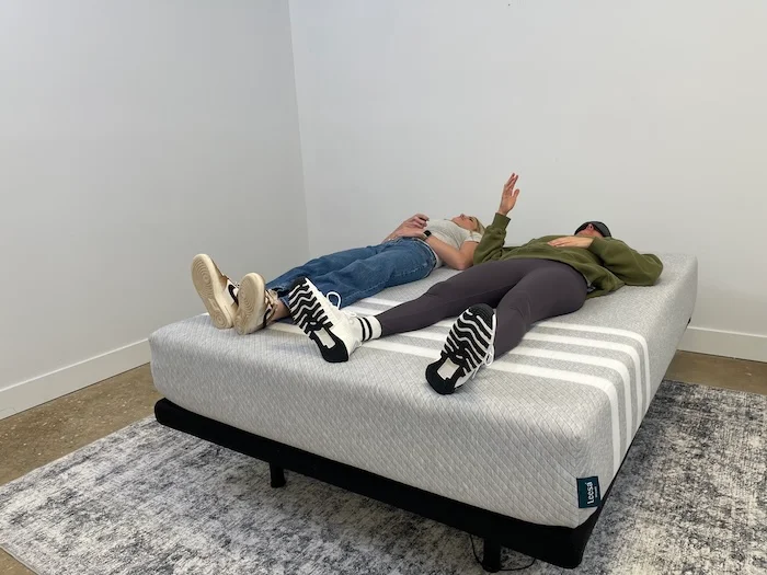Two women lying next to each other on the Leesa Original mattress. One of them is wearing a blindfold and using their hand to signal how much they can feel the other one's movement.