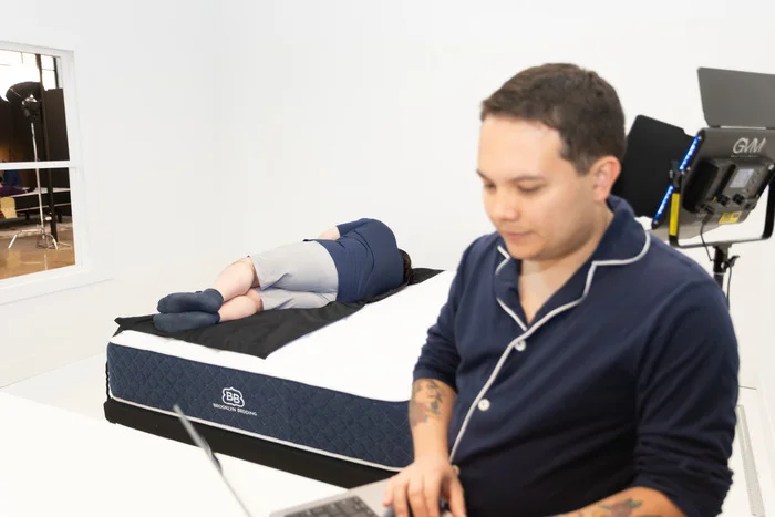 A man holding a laptop while another man behind him rests on a pressure mat on top of the Brooklyn Bedding Signature Hybrid mattress. The man with the laptop is getting a read on the mattress's pressure buildup on the tester's body.