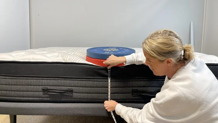 A woman kneeling next to the Nolah Evolution mattress while it has a stack of weights on it. She's measuring the bed's sinkage.