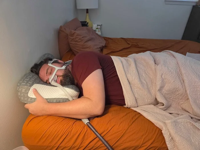 A man sleeping on his side on the Elviros pillow. He's wearing a sleep apnea mask as he dozes.
