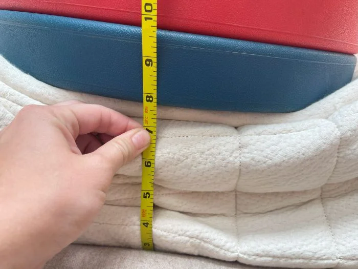 A stack of weight plates sit on top of the Avocado Luxury Organic mattress. A hand is measuring the sinkage with a tape measure.