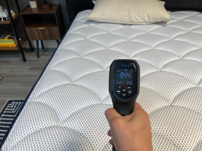 A closeup of someone using a thermal gun to take the temperature of the DreamCloud Premier Hybrid after someone has rested on it for 5 minutes. The screen reads 84.4 degrees.