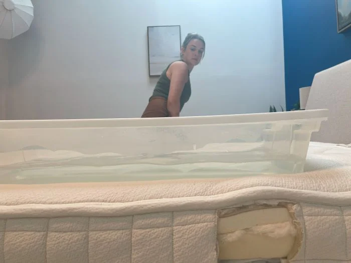 A woman sits on an Avocado Organic mattress next to a bin full of water.