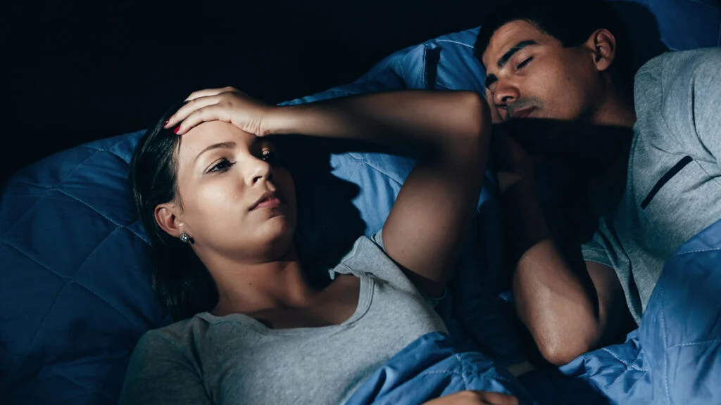 A woman placing a hand on her forehead while she tries to fall asleep next to her partner.