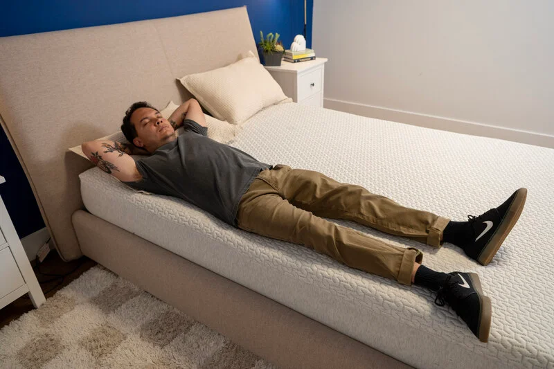 Kenny lies down near the very edge of the bed to see how much it sinks