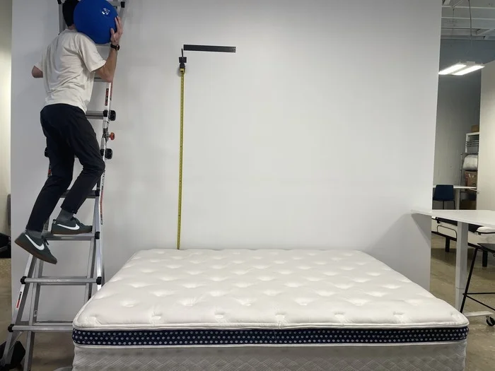 A man climbing a ladder with a large blue ball. He's about to drop it on the WinkBed to test the mattress's bounce.