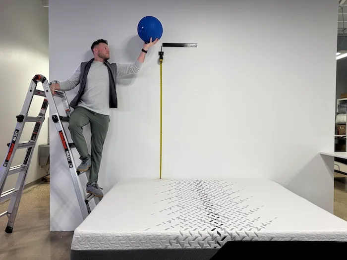 A man stands on a ladder and holds a 25-pound steel ball over the Nectar Luxe mattress.