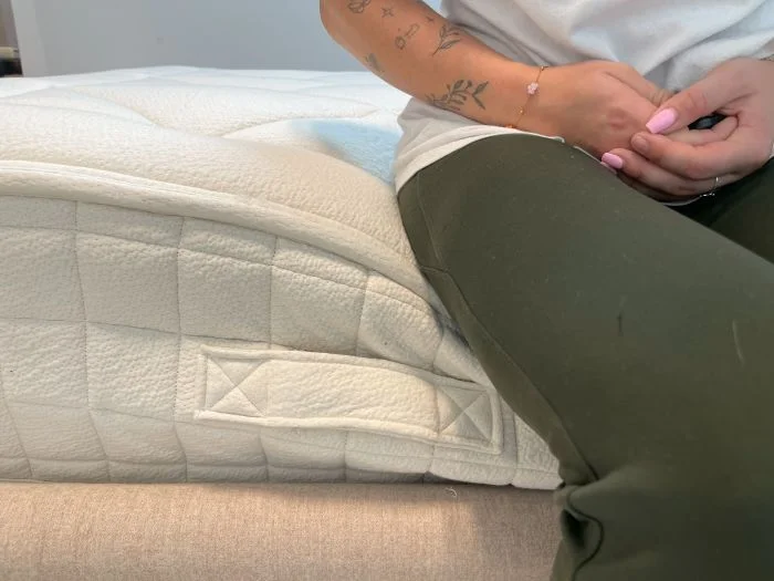 A closeup of the side of the Avocado Luxury Organic mattress. A woman is sitting on it and causing some sinkage to the layers.