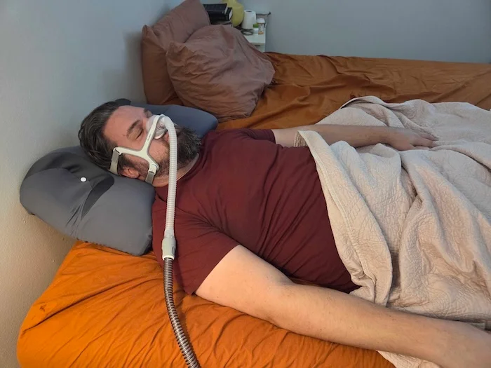 A man sleeping on his back on the Ikstar pillow. He's wearing a sleep apnea mask as he dozes.