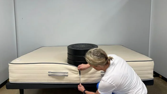 A woman kneeling next to a Nolah Natural mattress. A stack of weight sits on top of the bed and she's measuring the sinkage with a tape measure.