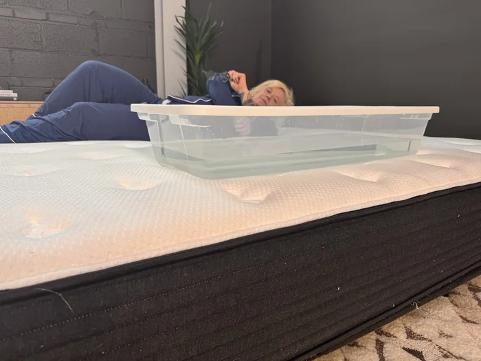 A woman lying on the Plank Firm mattress next to a plastic bin full of water. She's rolling around to test out the bed's motion isolation.