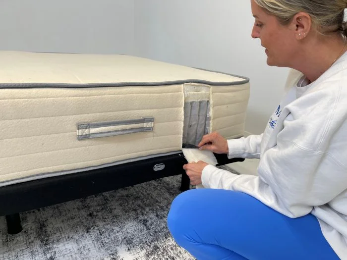 A woman kneeling next do a Nolah Natural mattress, peeling a piece of fabric off the side, and looking at its construction.