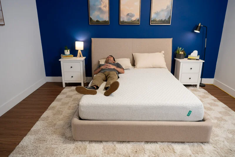 Kenny lies down near the very edge of the bed to see how much it sinks