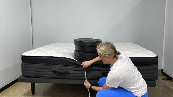 A woman kneeling next to the Nolah Evolution mattress while it has a stack of weights on it. She's measuring the bed's sinkage.