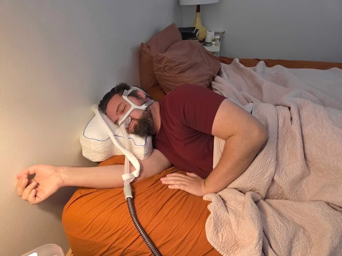 A man sleeping on his side on the Contour pillow. He's wearing a sleep apnea mask as he dozes.