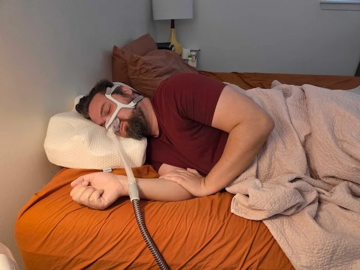A man sleeping on his side on the Lunderg pillow. He's wearing a sleep apnea mask as he dozes.