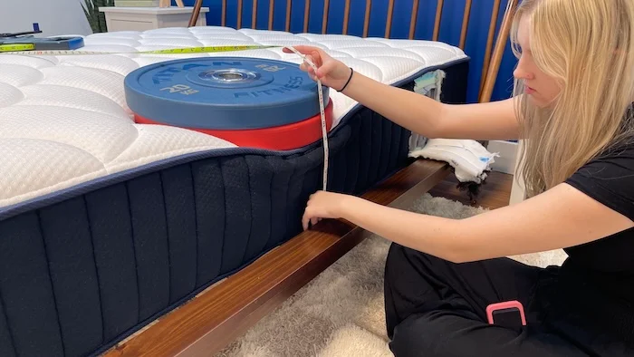 A woman kneeling next to the DreamCloud mattress. A stack of weights rests on the bed and the woman is measuring the sinkage with a tape measure.