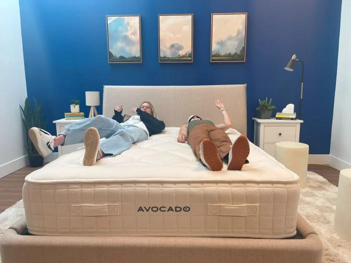 One woman lies on an Avocado Luxury Organic mattress while another one rolls around next to her.