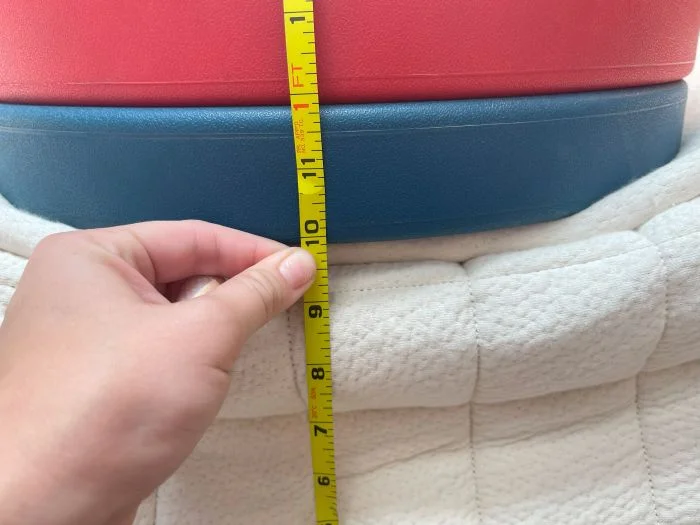 A stack of weight plates sit on top of the Avocado Luxury Organic mattress. A hand is measuring the sinkage with a tape measure.