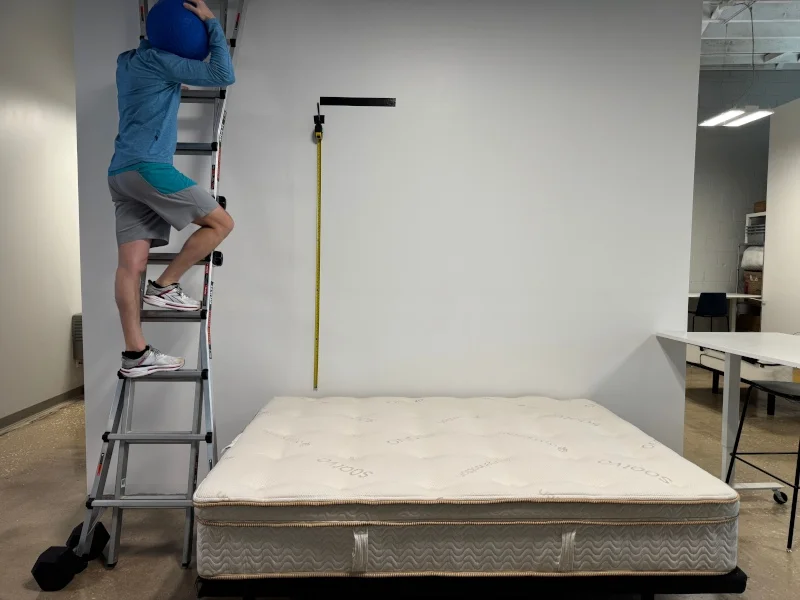 A person climbs a ladder to drop a medicine ball on a Saatva Latex Hybrid.