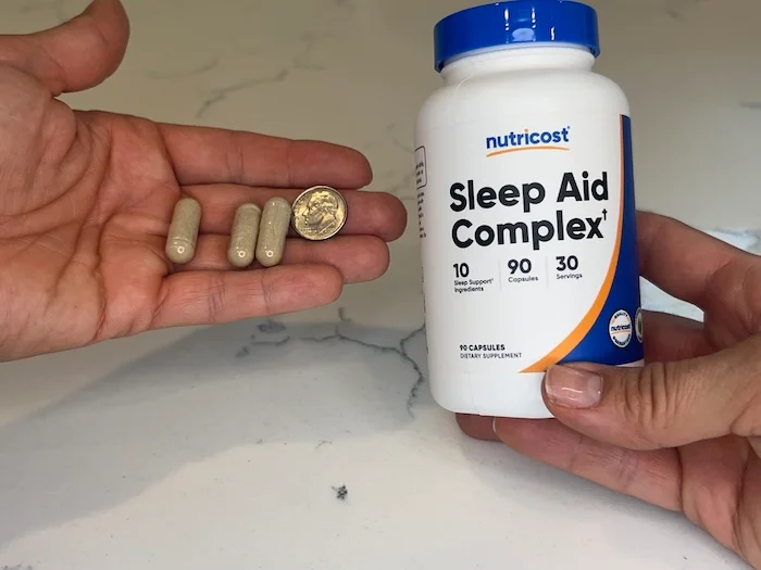 A hand holds Nutricost Sleep Aid Complex capsules next to a dime for size comparison.