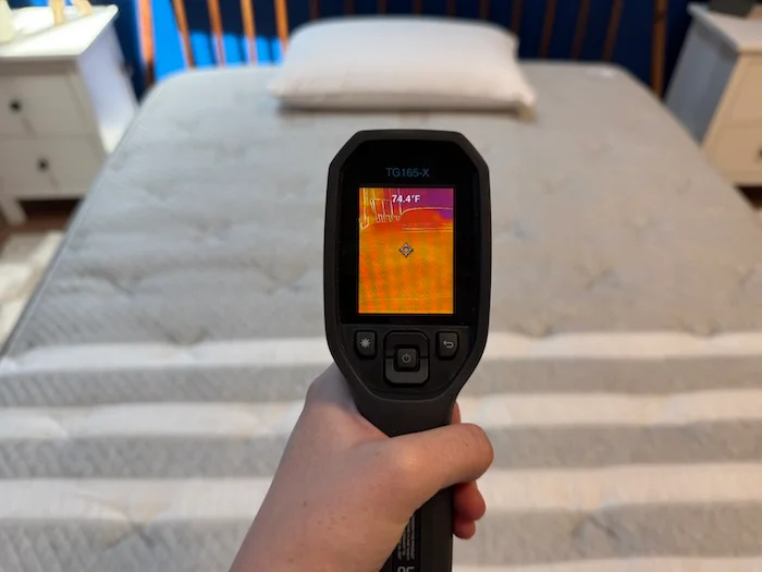 A closeup of a hand holding a thermal gun and using it to read the neutral temperature of a mattress. The screen says 74.4 degrees.