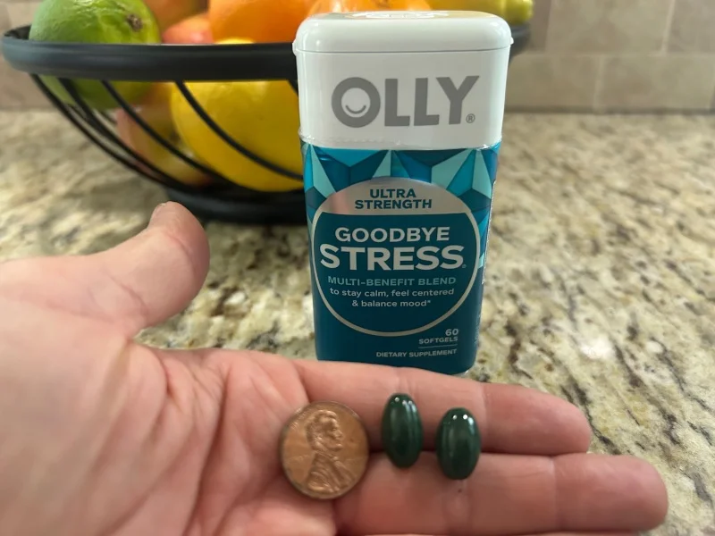 A hand holding some Olly Goodbye Stress gelcaps next to a penny for size comparison.