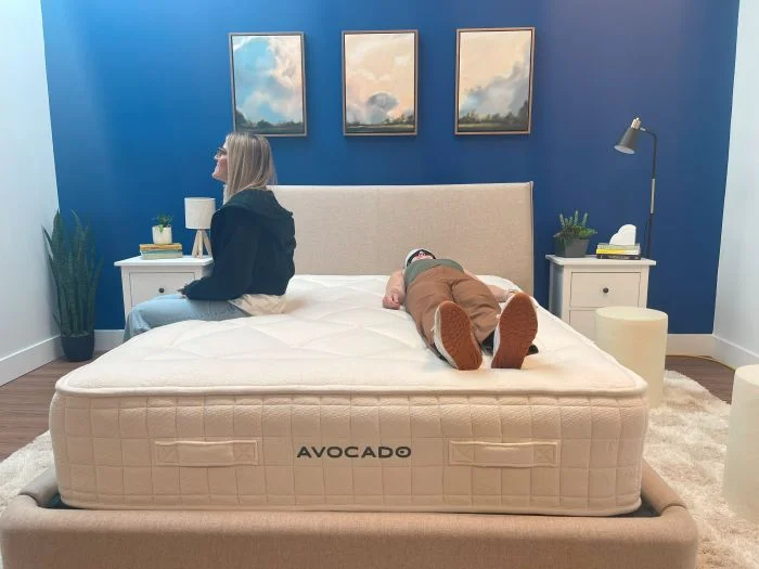 One woman sits on the edge of an Avocado Luxury Organic mattress while the other lies next to her with an eye mask on.