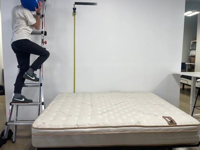 A many climbing a ladder next to the Saatva mattress with a medicine ball in his hand. He's about to drop it on the Saatva to test the bed's bounce.