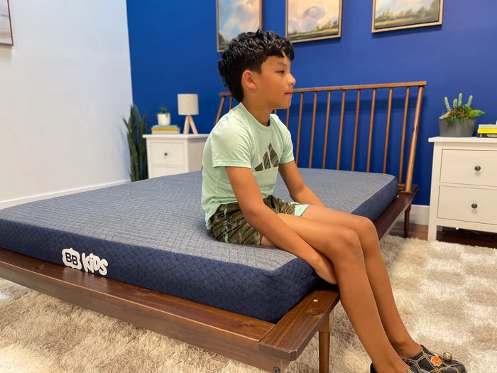 A 10-year-old boy sitting on the edge of the BB Kids mattress to test out its edge support.
