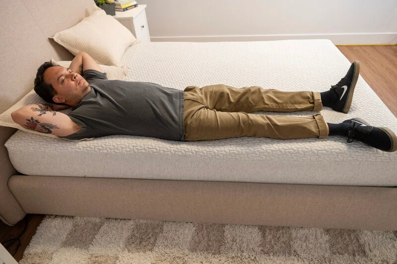 Kenny lies down near the very edge of the bed to see how much it sinks