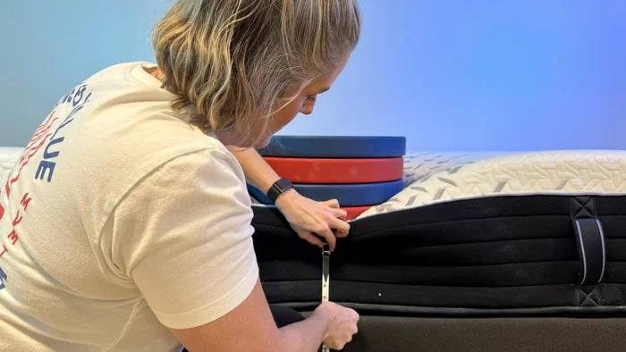 A woman kneeling next to the Nectar Luxe Hybrid mattress, which has a stack of weights on it. She's measuring the sinkage from the weights with a tape measure.