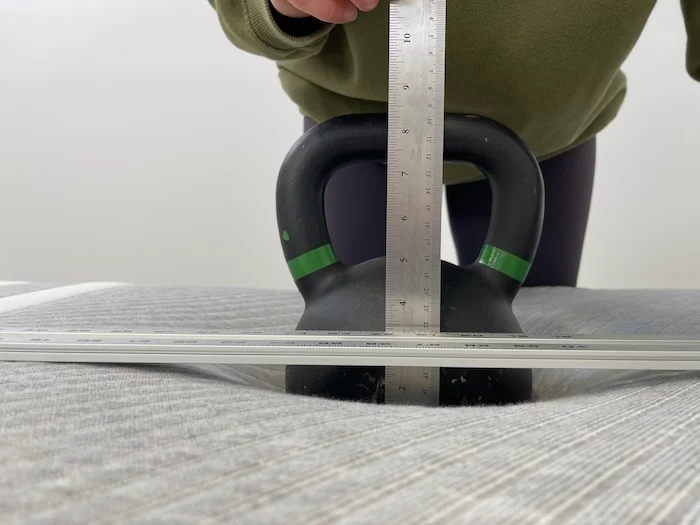 A kettlebell resting on a Leesa mattress. A woman is using a ruler to measure the sinkage.