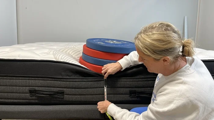 A woman kneeling next to the Nolah Evolution mattress while it has a stack of weights on it. She's measuring the bed's sinkage.