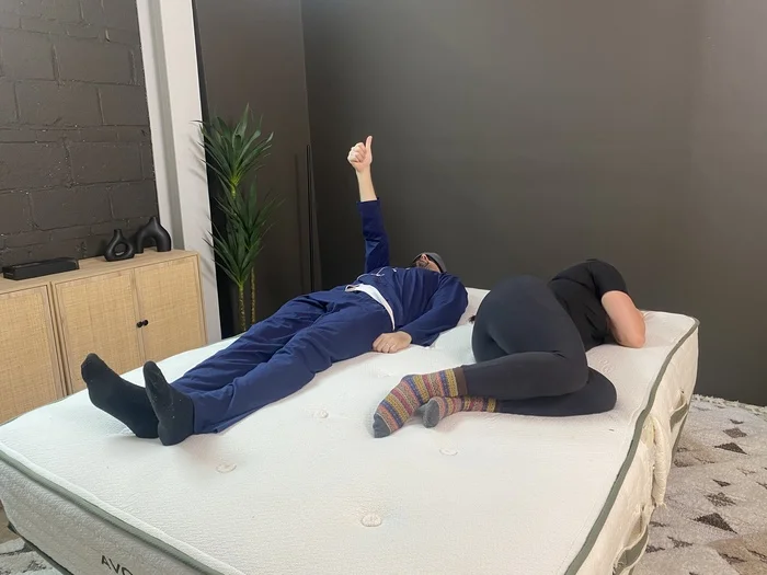 A man and a woman lying next to each other on the Avocado Green mattress. The man is wearing a sleep mask and giving a thumbs up, indicating that he can feel the woman moving around.