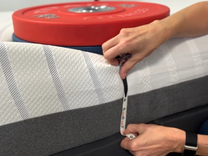A stack of weights resting on the BodiPedic 12" mattress. A woman kneels next to it, measuring the sinkage.