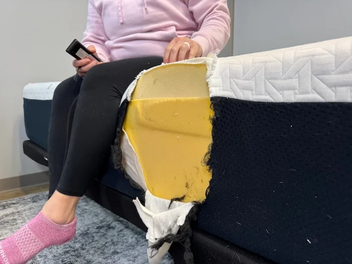 A woman sitting on the edge of the Nectar Luxe Memory Foam mattress. The side has been cut out, revealing the inner layers of the bed.