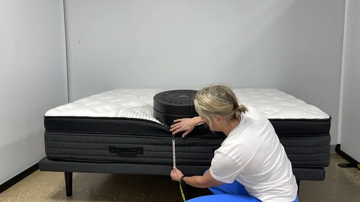 A woman kneeling next to the Nolah Evolution mattress while it has a stack of weights on it. She's measuring the bed's sinkage.