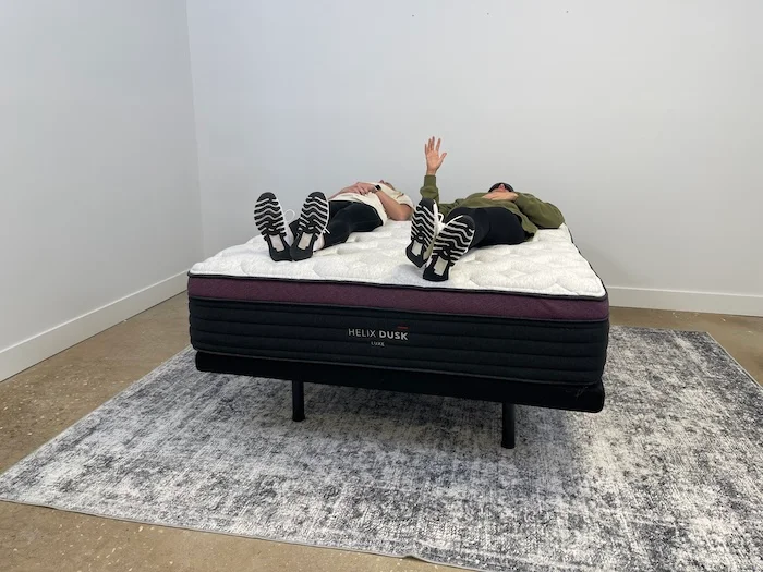 Two women lying side by side on the Helix Dusk Luxe mattress. One of them is wearing a sleep mask and raising her hand to indicate that she can feel the movement of her partner.