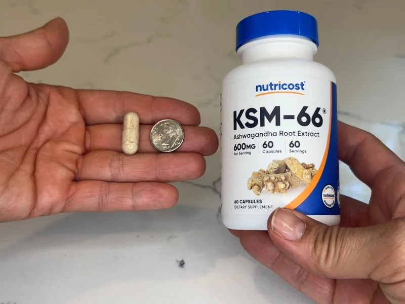 A hand holding a Nutricost KSM-66 capsule next to a dimefor size comparison.