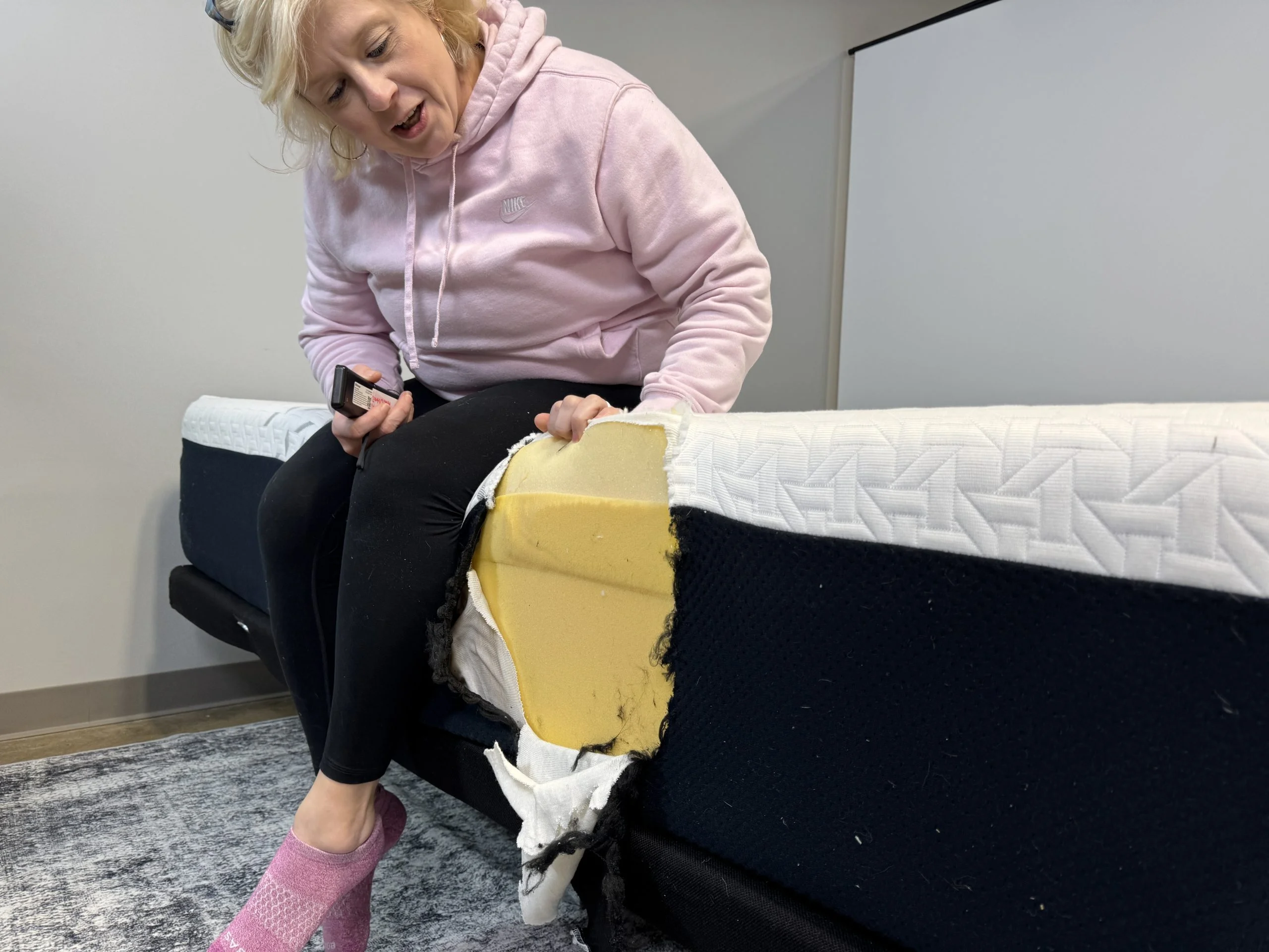 A person sits on the edge of a mattress, looking at a the layers where the mattress has been cut open.